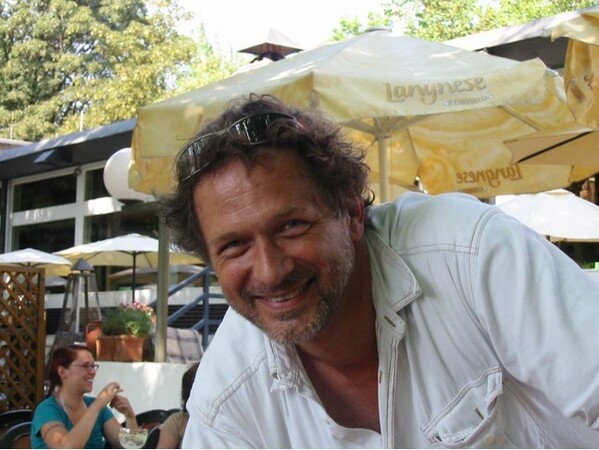 Ein Mann mit Sonnenbrille sitzt lachend im sonnigen Biergarten unter gelben Sonnenschirmen.